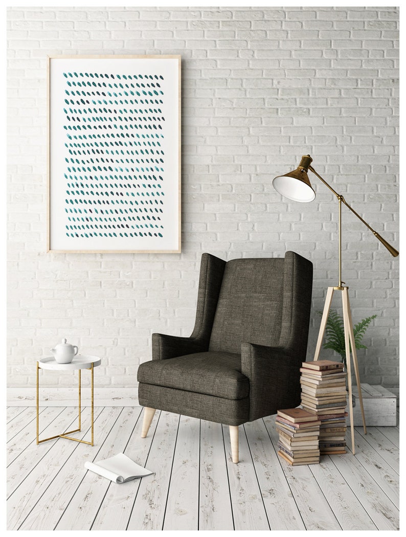 May include: A framed abstract art print with a pattern of teal and dark blue brushstrokes hangs on a white brick wall. A gray armchair with wooden legs sits below the artwork. A small side table and a floor lamp are also in the room.