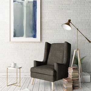 May include: An interior scene featuring a framed abstract watercolor painting in shades of blue and gray. A dark gray wingback chair with light wood legs sits near a white brick wall. A side table with a gold frame and a white teapot are also present.