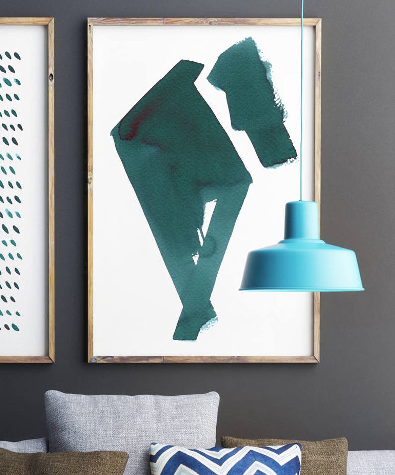 May include: Two framed abstract art prints with teal and dark teal watercolor designs. A light blue pendant lamp hangs in front of the artwork. The frames are made of light brown wood. The background is a dark gray wall.