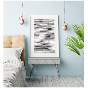 May include: A framed abstract art print featuring horizontal black lines on a white background. The artwork is displayed on a gray nightstand with silver legs, next to a bed with a beige headboard. Copper-colored pendant lights hang above.