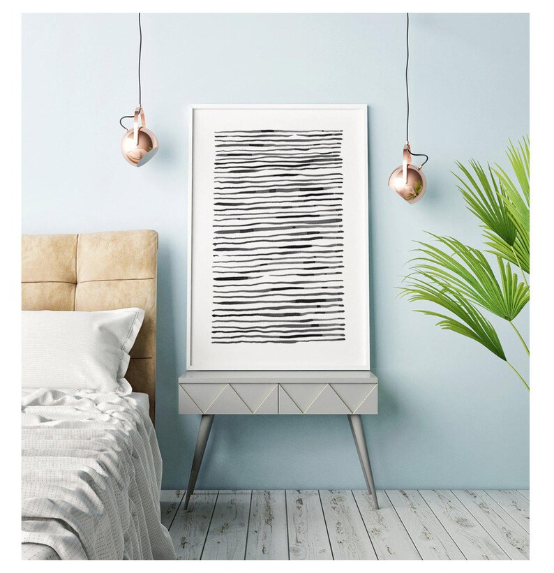 May include: A framed abstract art print featuring horizontal black lines on a white background. The artwork is displayed on a gray nightstand with silver legs, next to a bed with a beige headboard. Copper-colored pendant lights hang above.