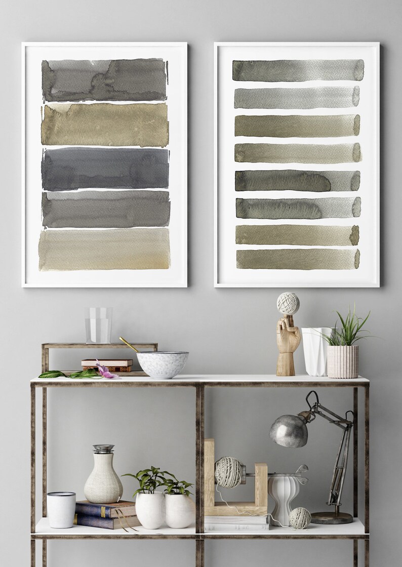 May include: Two framed watercolor art prints. Each print features horizontal brushstrokes in shades of gray, brown, and beige. The artwork is displayed on a white shelf with decorative items, including a lamp and plants.