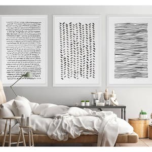 May include: Three framed abstract art prints. The prints feature black and white designs: a pattern of small dots, a vertical arrangement of leaf-like shapes, and horizontal lines. The artwork is displayed above a bed with white bedding.