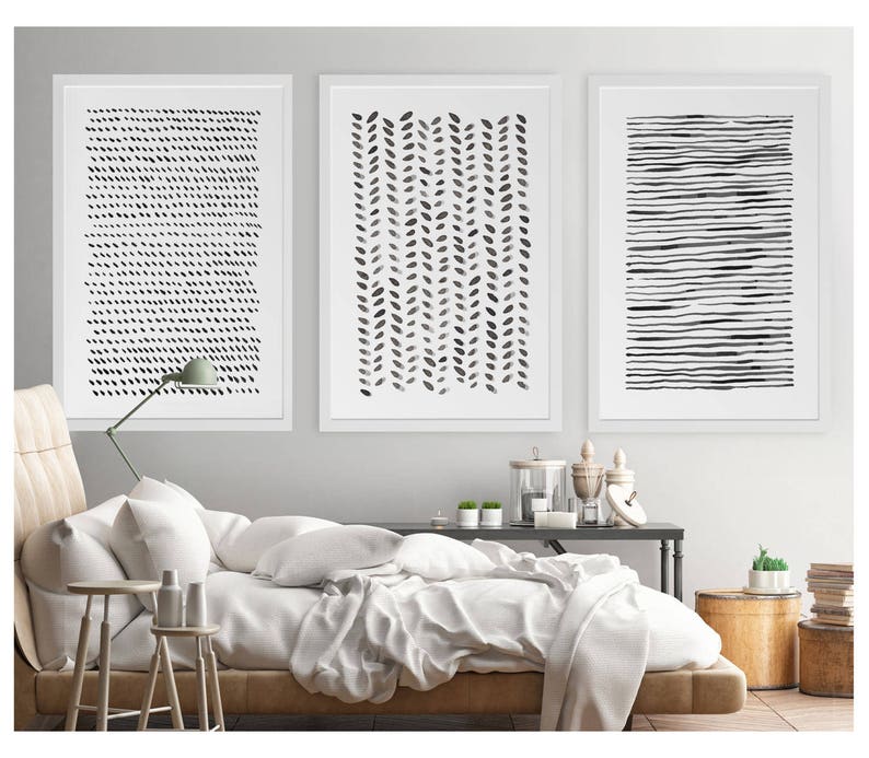 May include: Three framed abstract art prints. The prints feature black and white designs: a pattern of small dots, a vertical arrangement of leaf-like shapes, and horizontal lines. The artwork is displayed above a bed with white bedding.