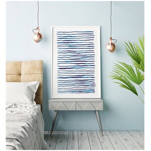 May include: A framed art print featuring horizontal blue lines of varying shades. The artwork is displayed on a gray nightstand with silver legs, next to a bed with a beige headboard. The room has a light blue wall and a copper-colored pendant light.