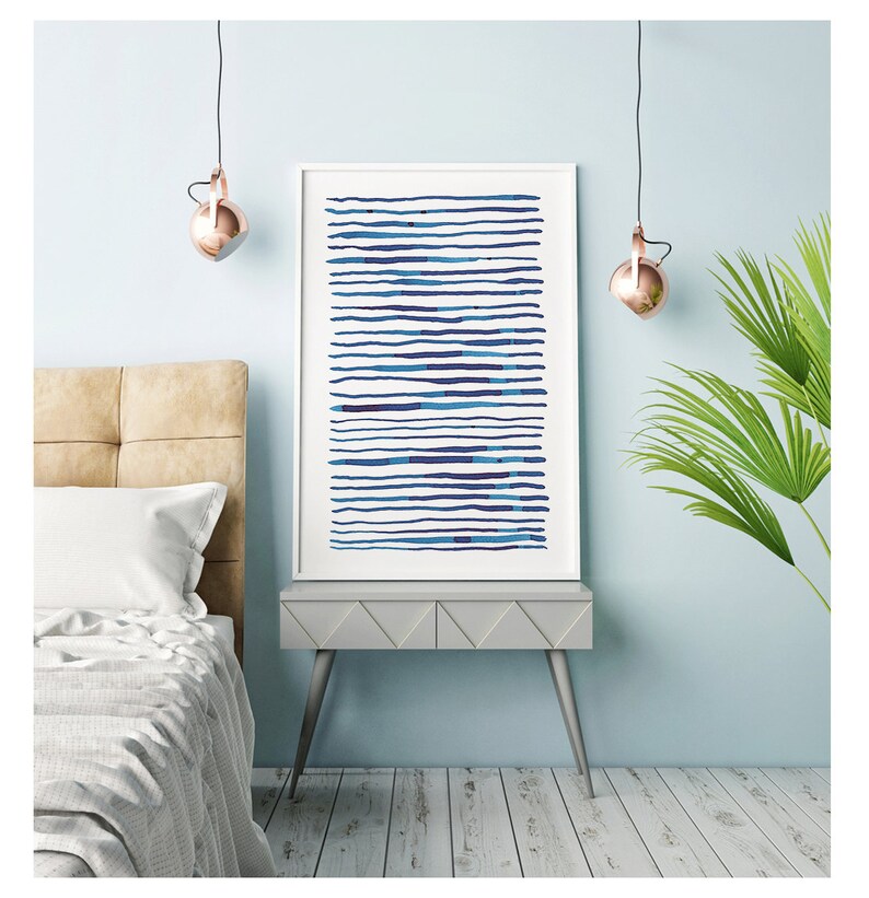 May include: A framed art print featuring horizontal blue lines of varying shades. The artwork is displayed on a gray nightstand with silver legs, next to a bed with a beige headboard. The room has a light blue wall and a copper-colored pendant light.