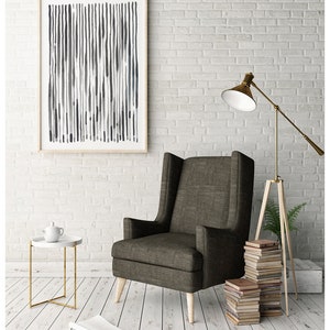 May include: A framed abstract art print with vertical lines in shades of gray and blue hangs on a white brick wall. A brown upholstered armchair with light wood legs sits below the artwork. A gold floor lamp and a small side table complete the room.
