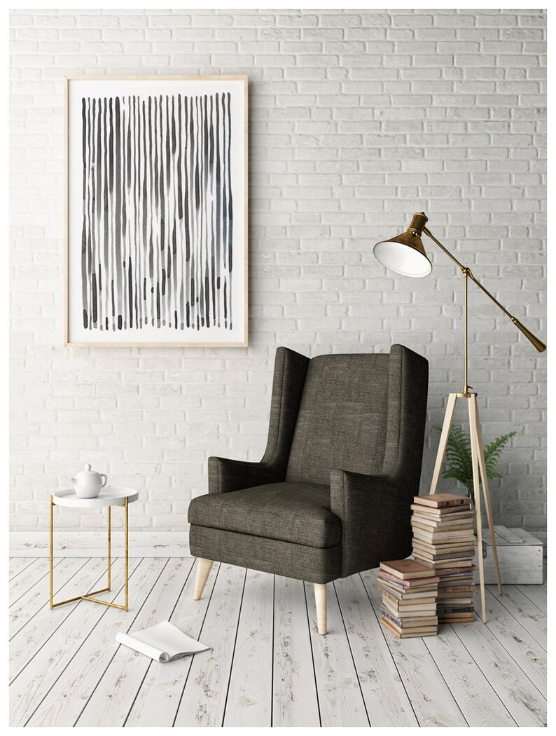 May include: A framed abstract art print with vertical lines in shades of gray and blue hangs on a white brick wall. A brown upholstered armchair with light wood legs sits below the artwork. A gold floor lamp and a small side table complete the room.