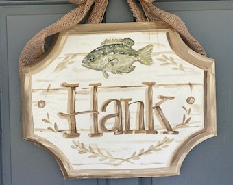 May include: A wooden sign with a beige background and brown trim. The sign has a painted image of a fish and the name "Hank" in brown lettering. The sign is hanging from a door with a burlap bow.