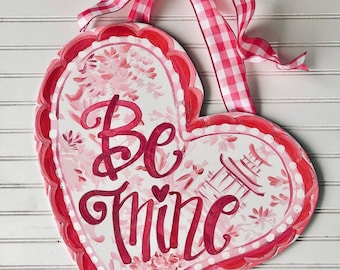 May include: A white heart-shaped wooden sign with a pink and white floral pattern. The sign says "Be Mine" in pink script.