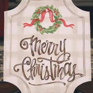May include: A cream-colored sign with a decorative border and the words "Merry Christmas" in brown script. A green wreath with red berries and a red ribbon is painted above the text. The sign is supported by a black stand.