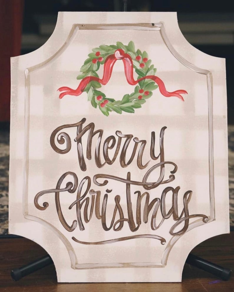 May include: A cream-colored sign with a decorative border and the words "Merry Christmas" in brown script. A green wreath with red berries and a red ribbon is painted above the text. The sign is supported by a black stand.