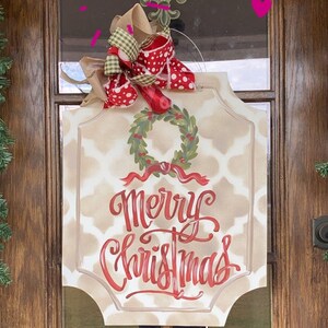 May include: A rectangular Christmas sign with a beige patterned background and the words "Merry Christmas" in red script. A green wreath with red berries and a red ribbon are above the text. The sign is topped with a decorative bow.