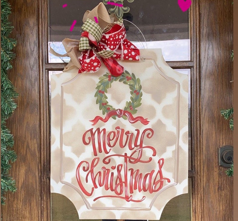 May include: A rectangular Christmas sign with a beige patterned background and the words "Merry Christmas" in red script. A green wreath with red berries and a red ribbon are above the text. The sign is topped with a decorative bow.