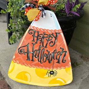 May include: A wooden candy corn shaped door hanger with the words "Happy Halloween" painted in black. The candy corn is decorated with orange, yellow, and white paint and has a black spider hanging from a string.