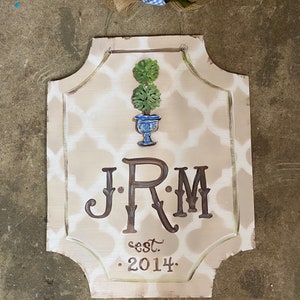 May include: A light brown wooden sign with a white and brown geometric pattern. The sign features a painted image of a topiary in a blue and white planter. The sign also includes the initials "J.R.M." and the text "est. 2014."
