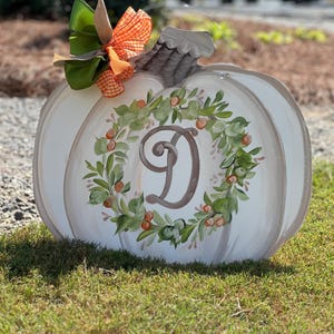 May include: A white pumpkin-shaped wooden cutout featuring a hand-painted green wreath with orange berries surrounding a large, dark brown script letter D in the center. The pumpkin has a grey stem and a decorative bow made of green and orange ribbons.