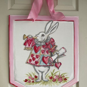 May include: A white rabbit wearing a red and blue heart-shaped vest and holding a scroll and a trumpet. The rabbit is standing on a green patch of grass with pink flowers. The image is painted on a white wooden sign with a pink border.
