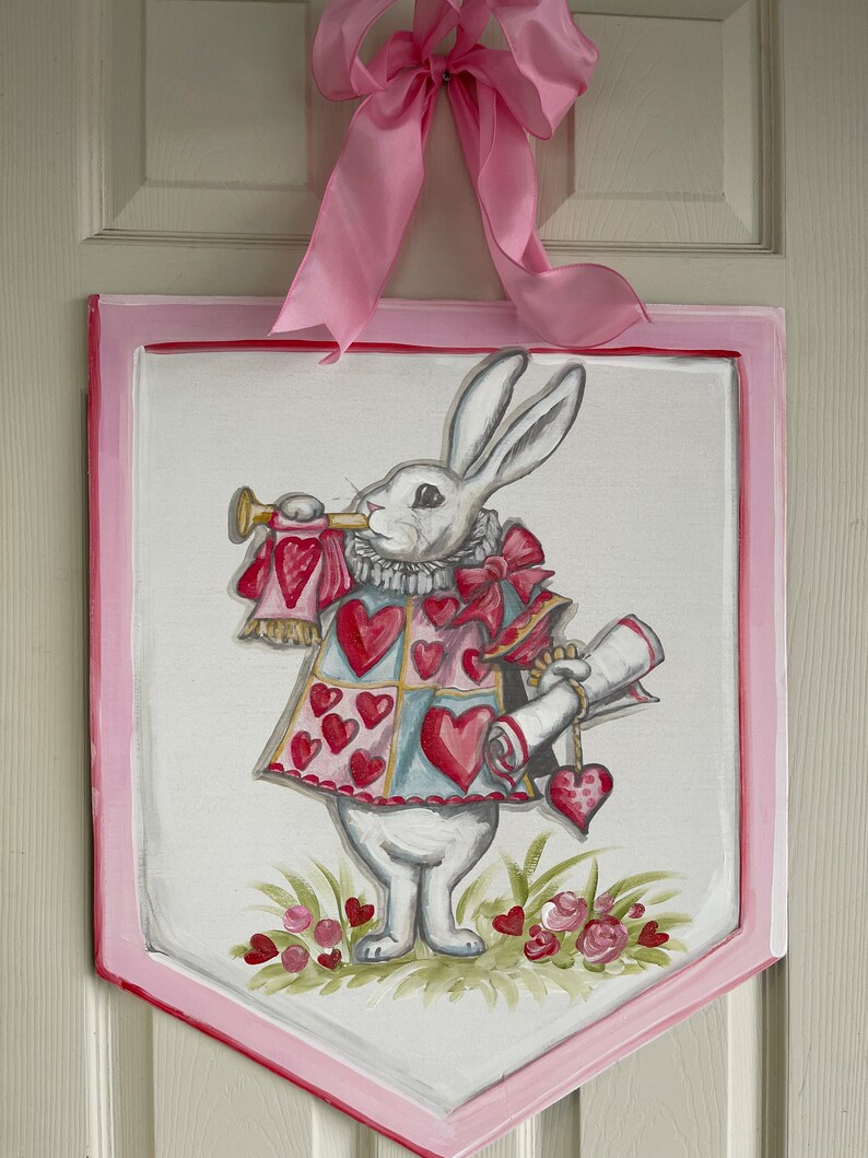May include: A white rabbit wearing a red and blue heart-shaped vest and holding a scroll and a trumpet. The rabbit is standing on a green patch of grass with pink flowers. The image is painted on a white wooden sign with a pink border.