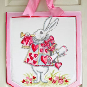 May include: A white rabbit wearing a red and blue heart-patterned vest and holding a scroll and a trumpet. The rabbit is painted on a pink and white wooden sign with a scalloped edge.