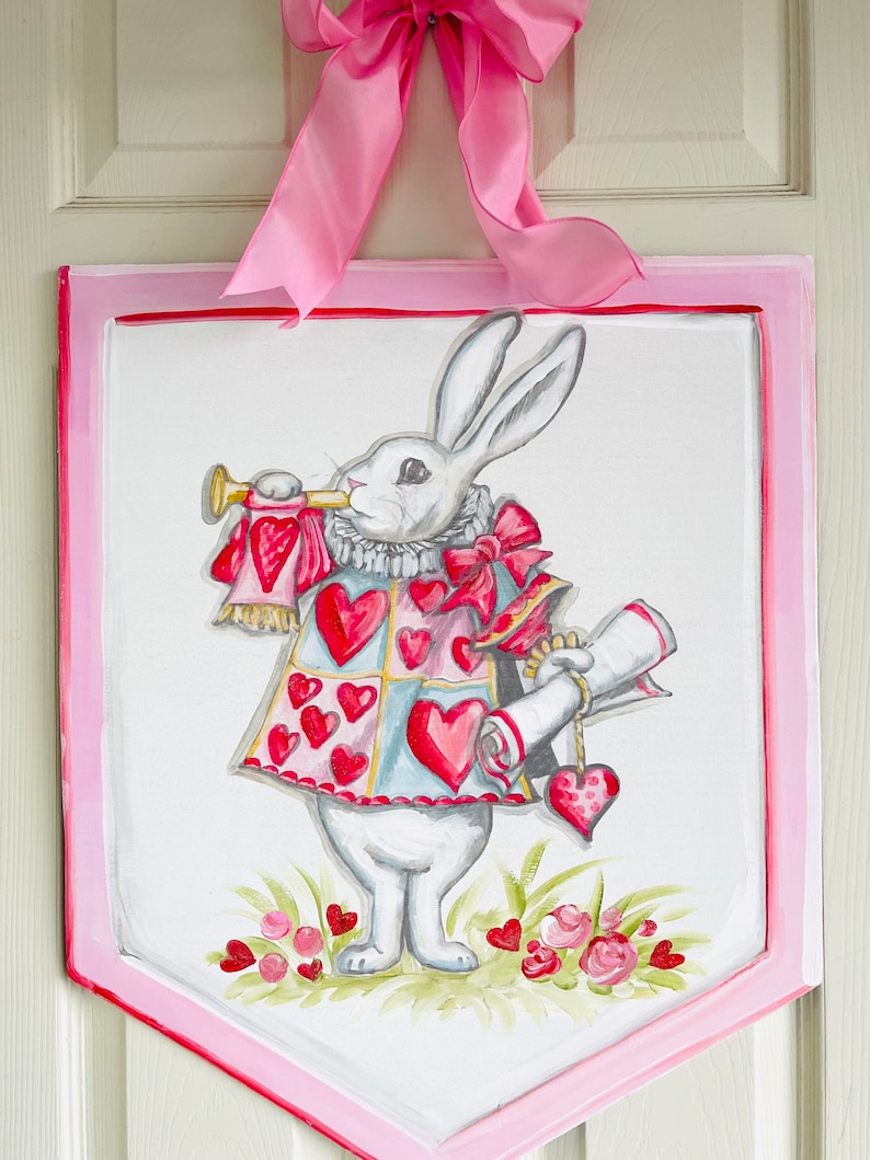 May include: A white rabbit wearing a red and blue heart-patterned vest and holding a scroll and a trumpet. The rabbit is painted on a pink and white wooden sign with a scalloped edge.