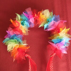 May include: A colorful rainbow-themed headband with a semi-circular design. The headband is made of layered tulle in various colors, including red, orange, yellow, green, blue, and purple. Red ribbons with white polka dots and solid red ribbons hang from the ends.