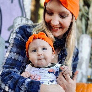 May include: A woman wearing a bright orange beanie with a bow holds a baby wearing a white onesie with a pumpkin and bat print. The baby is also wearing a bright orange headband with a bow. The woman and baby are smiling at the camera.