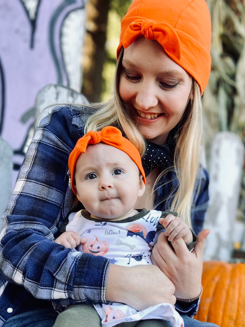 May include: A woman wearing a bright orange beanie with a bow holds a baby wearing a white onesie with a pumpkin and bat print. The baby is also wearing a bright orange headband with a bow. The woman and baby are smiling at the camera.