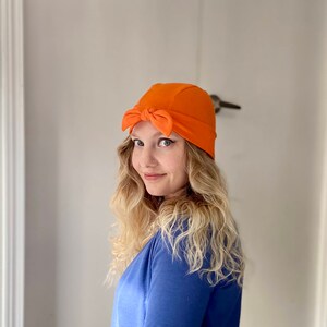May include: A woman with long blonde hair is wearing a bright orange turban-style hat with a bow on top. She is wearing a blue shirt and looking to the side.