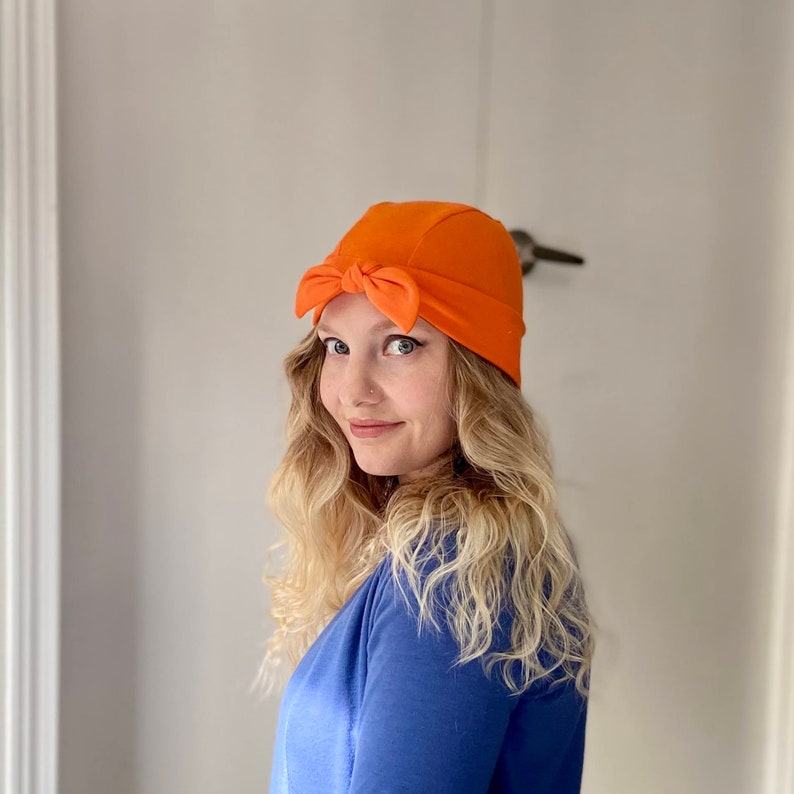 May include: A woman with long blonde hair is wearing a bright orange turban-style hat with a bow on top. She is wearing a blue shirt and looking to the side.