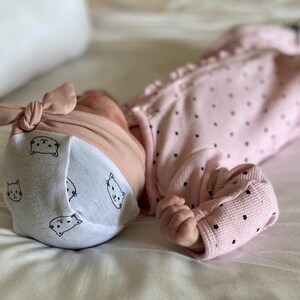 May include: A pink and white baby hat with a knotted bow on top. The hat has a white background with black cat faces printed on it. The hat is on a white surface.