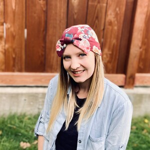 May include: A woman with blonde hair is wearing a floral patterned headband with a bow on top. She is wearing a light blue button-down shirt over a black shirt.