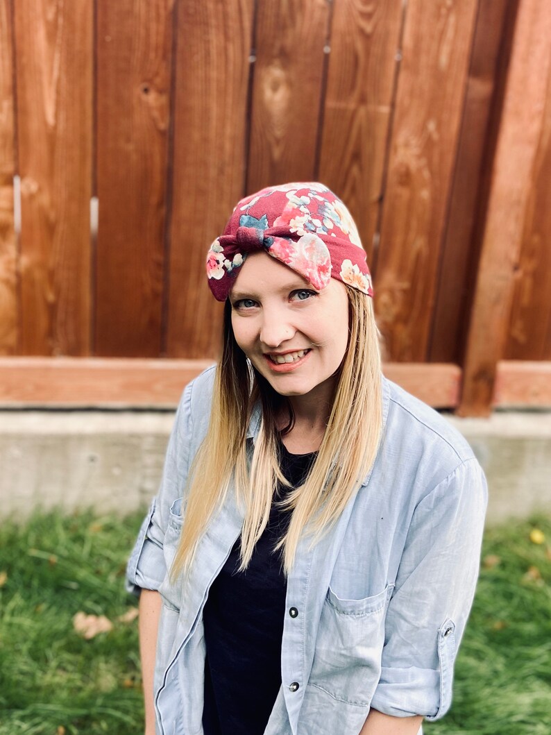 May include: A woman with blonde hair is wearing a floral patterned headband with a bow on top. She is wearing a light blue button-down shirt over a black shirt.