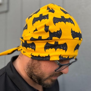 May include: A yellow fabric bandana with a black bat pattern. The bandana is tied around a person's head.