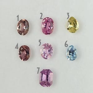 May include: An assortment of seven gemstones in various shapes and colors. The gems include oval, pear, and emerald cuts, with hues ranging from pink and purple to yellow and blue. The gemstones are displayed on a white surface.