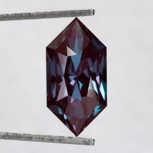 Puede incluir: Una gema facetada con forma hexagonal, sujeta con pinzas metálicas. La gema muestra un degradado de color, pasando del borgoña oscuro a tonos turquesa y púrpura. El corte de la piedra crea múltiples superficies reflectantes.