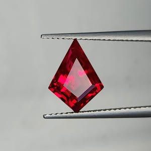 Lab grown pigeon blood red ruby in precision kite cut, held upright in gem tweezers against a neutral studio background. The stone shows sharp step facets, flawless clarity, and intense red saturation from edge to center. AAA grade synthetic ruby with expert Thai polishing, ready for a custom engagement ring, pendant, or statement earring design.