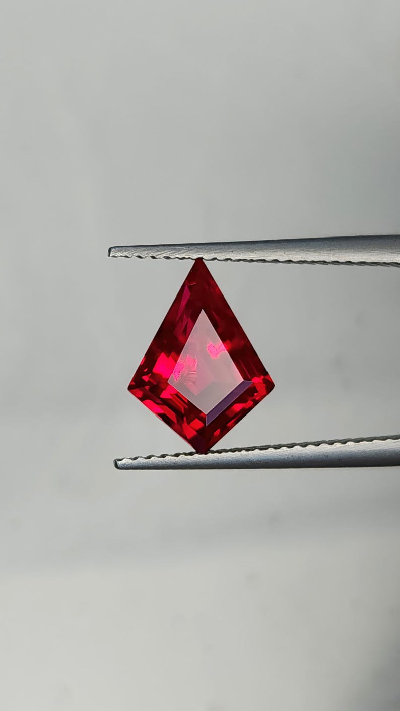 Lab grown pigeon blood red ruby in precision kite cut, held upright in gem tweezers against a neutral studio background. The stone shows sharp step facets, flawless clarity, and intense red saturation from edge to center. AAA grade synthetic ruby with expert Thai polishing, ready for a custom engagement ring, pendant, or statement earring design.