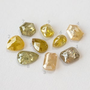 May include: Nine loose, natural diamonds in various shapes and shades of yellow, brown, and clear. The diamonds are scattered on a white surface, showcasing their unique cuts and colors. The shapes include pear, hexagon, and rectangular cuts.