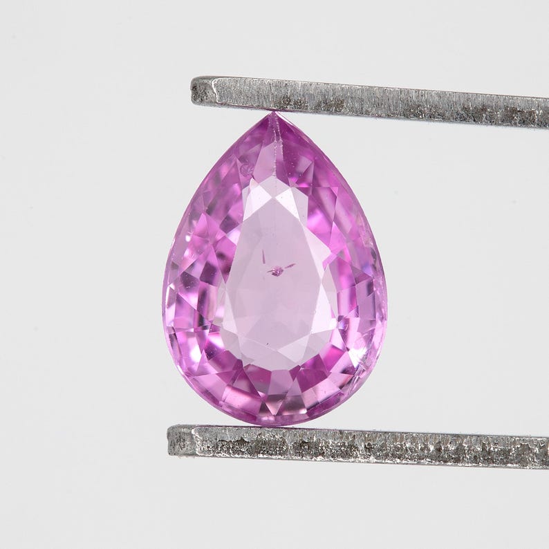 Face up view of a pear cut unheated natural sapphire in tweezers, vivid orchid pink color, bright reflections across the crown facets, Madagascar loose gemstone 0.75 ct, approx 6.9x5.0mm, ideal for a solitaire ring center stone.
