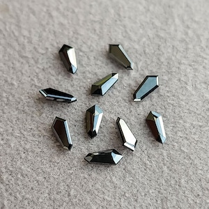 May include: A collection of ten dark gray, coffin-shaped crystals scattered on a light gray surface. Each crystal has multiple facets, reflecting light and creating a shimmering effect. The crystals are uniform in size and shape.