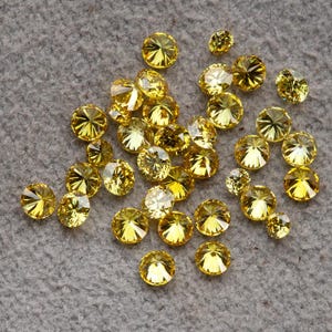 Puede incluir: Un conjunto de gemas amarillas redondas y facetadas dispersas sobre una superficie gris. Las gemas varían en tamaño, con múltiples facetas que capturan la luz, creando un efecto brillante. Las gemas son de un color amarillo vibrante y soleado.