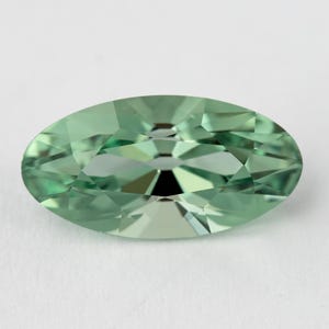 Puede incluir: Una piedra preciosa de corte ovalado con un tono verde menta claro y una superficie facetada. La piedra presenta una disposición simétrica de facetas triangulares y trapezoidales que crean un patrón geométrico reflectante.