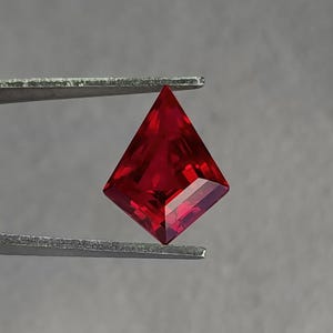 Bright pigeon blood red lab grown ruby displayed in gem tweezers under neutral lighting. The modern kite / shield cut gives a strong geometric outline for custom engagement rings and avant-garde pendants. Ultra-clean clarity and reflective facet work show the premium cutting standard. This loose synthetic ruby is a ready-to-set centerpiece.