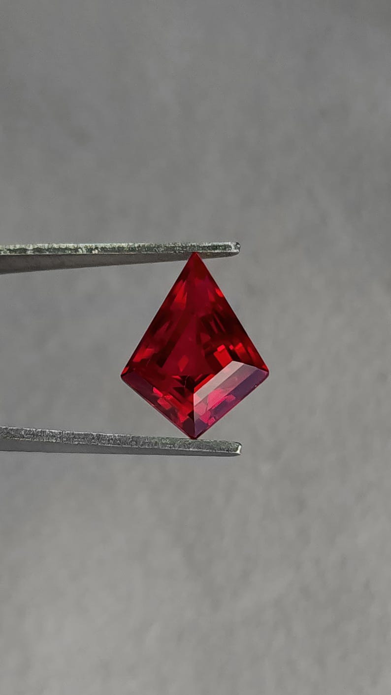Bright pigeon blood red lab grown ruby displayed in gem tweezers under neutral lighting. The modern kite / shield cut gives a strong geometric outline for custom engagement rings and avant-garde pendants. Ultra-clean clarity and reflective facet work show the premium cutting standard. This loose synthetic ruby is a ready-to-set centerpiece.