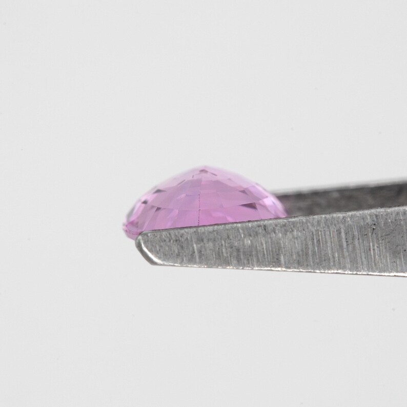 Side profile photo of the pear cut unheated natural sapphire in tweezers, vivid orchid pink tone visible along the girdle, smooth silhouette and depth view, Madagascar natural sapphire for a solitaire design.