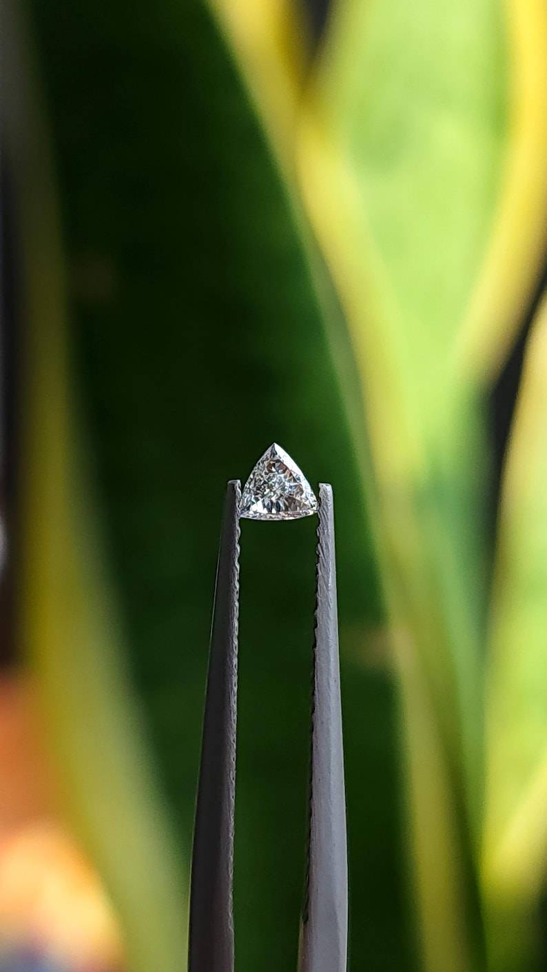 single 3x3mm trillion cut natural diamond held in tweezers side profile against green background, calibrated triangle stone showing crown and pavilion, bright natural daylight reflections, F G VVS VS side stone