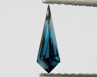 Puede incluir: Una piedra preciosa suelta en forma de cometa de color azul cerceta profundo con una superficie facetada. La piedra se sostiene verticalmente con unas pinzas de metal, mostrando sus extremos puntiagudos y corte geométrico.