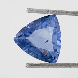 Op de afbeelding: Een driehoekige, gefacetteerde edelsteen met een levendige blauwe tint. De edelsteen wordt vastgehouden door een metalen pincet, wat de gepolijste oppervlakken en interne helderheid laat zien. De vorm is een gelijkzijdige driehoek, die licht en kleur reflecteert.