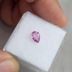Side angle of the 0.75ct pink sapphire, unheated and natural. The pear cut loose gemstone glows with vivid magenta rose hues, showing well-balanced symmetry and brilliance. Ideal for bespoke jeNatural sapphire placed on a white jewelry box pad held in hand, pear cut vivid orchid pink color, unheated Madagascar sapphire shown for true size and setting scale, suitable for custom jewelry.welry design, pendants, or gemstone collections.